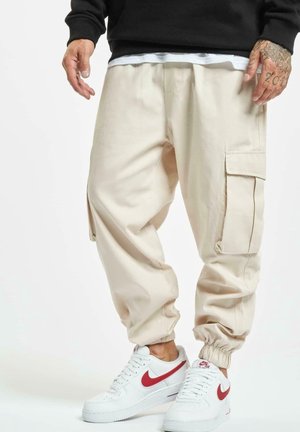 Personne portant un pantalon cargo beige avec des poignets élastiques, associé à des baskets blanches Nike Air avec des logos Swoosh rouges.