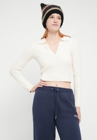 Hvid, kabelstrikket cropped sweater med V-hals og krave, kombineret med mørkeblå joggingbukser med broderet logo. Sort stribet hue.