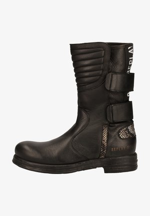 Mustassa nahkabootsissa on uritettu yksityiskohta, säädettävät hihnat, tekstuuriset akuutit ja litteä kumipohja. Kenkässä on hienovarainen käärmeennahkakuosi.