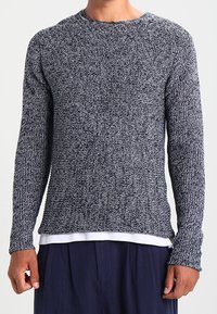 Marineblauer und grauer Strickpullover mit langen Ärmeln, rundem Halsausschnitt und strukturiertem Muster; getragen über einem weißen, mehrlagigen Hemd.