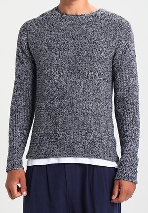 Marineblauer und grauer Strickpullover mit langen Ärmeln, rundem Halsausschnitt und strukturiertem Muster; getragen über einem weißen, mehrlagigen Hemd.