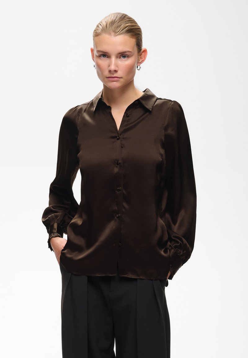 Jeune femme aux cheveux blonds coiffés en chignon lisse, portant un chemisier en satin marron foncé et un pantalon noir, debout les mains dans les poches.