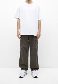 Person in einem schlichten weißen, übergroßen T-Shirt, dunkelgrünen Cargo-Hosen mit Seitentaschen und weißen Sneakers, der vor einem einfarbigen Hintergrund steht.