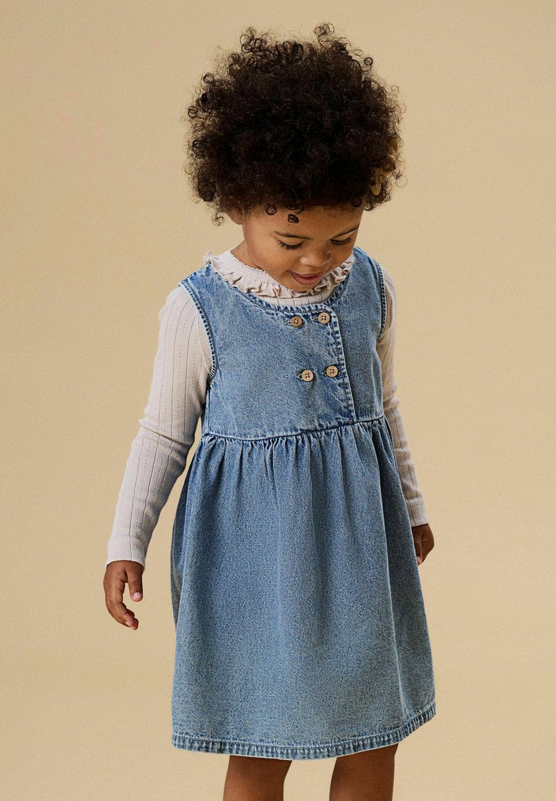 Zila džinsa kleita ar pogu detaļām, zvana formas svārkiem un savilktu vidukli, valkāta virs baltas garām piedurknēm izstrādātas virsdrēbes ar ribotu faktūru un ruļļotu apkakli.