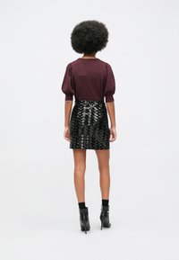 Nainen, jolla on kiharat hiukset, seisoo sel backs, päällä burgundinvärinen puffihihainen paita, musta säihkeä mini-hame, mustat sukat ja mustat korkokengät.