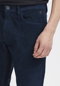 Sötétkék corduroy nadrág függőleges bordákkal, öt zsebes kialakítással és gombos záródással. A logó a front zsebben található. Puha tapintású.