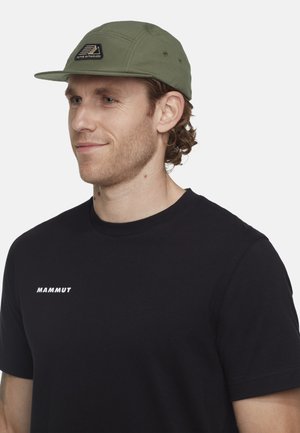 Mand med lys hud og krøllet brunt hår iført en olivengrøn kasket med "Alpine Enthusiasm" patch og en sort t-shirt med Mammut-logo.