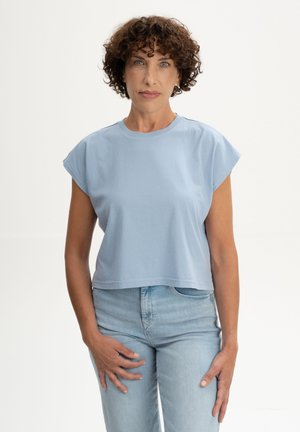 Femme aux cheveux courts et bouclés portant un haut sans manches bleu clair et un jean bleu clair, debout devant un fond blanc uni.