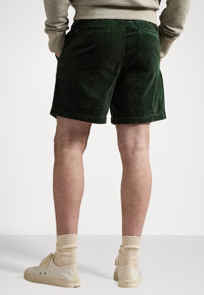 Pantaloni scurți din corduroy verde, de lungime medie până la coapsă, cu un brâu ce include bucle pentru curea și buzunare pe spate. Textură moale cu nervuri verticale.