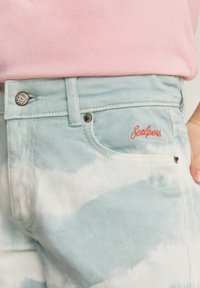 Jeans de mezclilla azul claro con un patrón de tie-dye, presentan un botón plateado, remaches negros y una marca bordada roja en el bolsillo trasero.