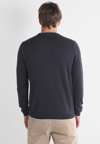 Homme portant un pull à manches longues gris foncé et un pantalon beige, vu de dos sur fond blanc uni.