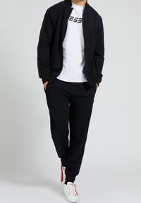 Veste bomber noire avec poignets côtelés, accompagnée d'un t-shirt blanc avec un logo noir et d'un pantalon de jogging noir. Baskets blanches avec des accents rouges.