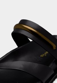 Κοντινό πλάνο μαύρου σανδαλιού με δύο λουριά, ένα από τα οποία διαθέτει λαμπερή μεταλλική χρυσή λεπτομέρεια.