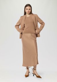 Beżowa marynarka dopasowana nałożona na pasujący sweter z dzianiny oraz satynową spódnicę midi. Brązowe kozaki na kostkę związane na sznurówki dopełniają stylizację gładką fakturą.