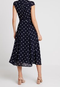 Granatowa sukienka w groszki z krótkimi rękawkami typu kapturek, dopasowaną talią, rozkloszowanym dołem i delikatną fakturą. Posiada pasek i sięga do kolan.