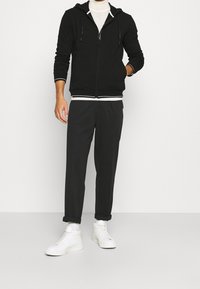 Felpa nera con zip e cordini, dettagli bianchi, abbinata a pantaloni jogger scuri e sneakers alte bianche. Texture liscia in tutto.