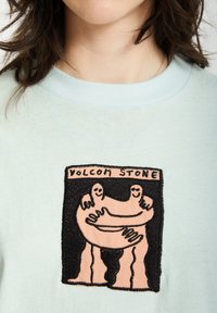 Hellblaues Baumwoll-T-Shirt mit einem schwarz-rosafarbenen gestickten Grafikdesign von zwei sich umarmenden Figuren, mit dem Text "VOLCOM STONE" darüber.