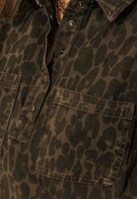 Chemise en velours côtelé marron présentant un imprimé léopard foncé, une fermeture boutonnée à l'avant et deux poches poitrine. Tissu texturé avec des détails de couture visibles.