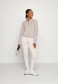 Svijetlosmeđi zip-zatvarač jakne s rebrastim manžetama, u kombinaciji s mekim, svijetlopinkastim trenirkama. Model drži konopac za preskakanje, odjeven u bijele atletske tenisice.