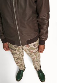 Man i mörkbrun läderjacka, beige kamouflagebyxor och gröna snörade kängor som står mot en vit bakgrund.