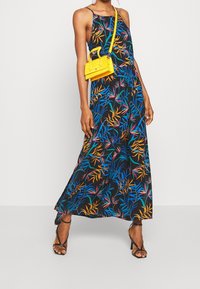 Nainen, jolla on yllään musta hihaton maksimekko, jossa on värikäs trooppinen lehtikuvio, mustat korkokengät ja pieni keltainen olkalaukku.