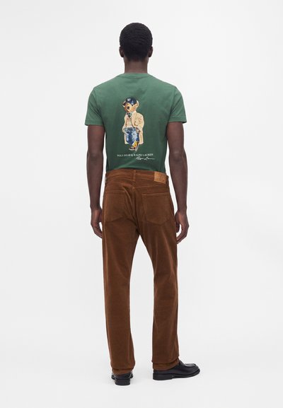 Uomo con t-shirt verde con grafica di un orso e pantaloni marroni, in piedi di spalle su sfondo bianco semplice.
