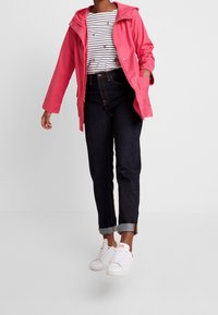 Rosa vattentät jacka med huva, bärs över en randig skjorta, kombinerad med mörka jeans och vita sneakers. Avslappnat outfit med uppvikt ärmar.