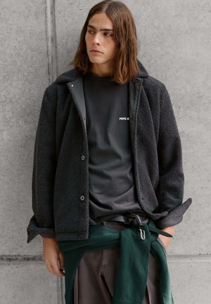 Junger Mann mit schulterlangem braunem Haar trägt eine schwarze Fleecejacke über einem schwarzen Pepe Jeans T-Shirt, mit einem grünen Pullover um die Taille gebunden.