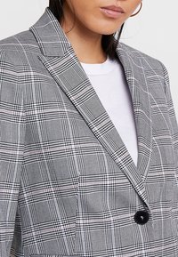 Kobieta ubrana w czarną, białą i różową kratkowaną marynarkę z jednym czarnym guzikiem na białej bluzce, widoczne złote kolczyki w kształcie kół.