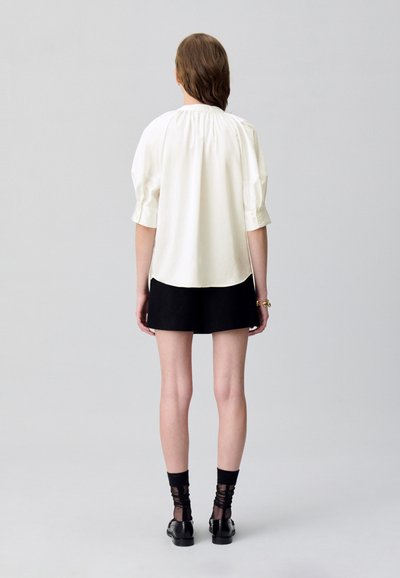 Blusa blanca con escote fruncido, mangas cortas de globo, combinada con una falda corta negra. La modelo lleva zapatos negros con calcetas negras texturizadas.