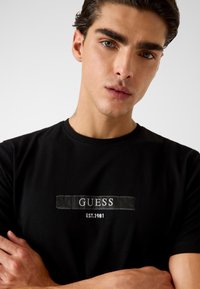Zwart katoenen T-shirt met een geborduurd "GUESS"-logo en "EST.1981" eronder, ronde halslijn en korte mouwen. Soepele textuur.