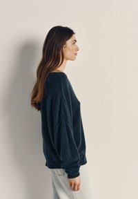 Mørkeblå oversized sweater med en bred halsudskæring, dropped skuldre og ribbede manchetter, parret med lyse denimjeans. Glat tekstur.