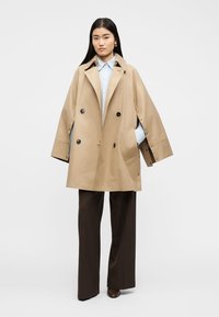 Νεαρή γυναίκα φοράει υπερμεγέθη μπεζ καμπαρντίνα, ανοιχτό μπλε πουκάμισο με ρίγες, σκούρα καφέ παντελόνια φαρδιάς γραμμής και καφέ παπούτσια μπροστά σε απλό φόντο.