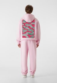 Rozā kapuce un atbilstošas sporta bikses. Kapucei aizmugurē ir spilgta grafiska apdruka zaļā un rozā krāsā. Audums izskatās mīksts un ar tekstūru.