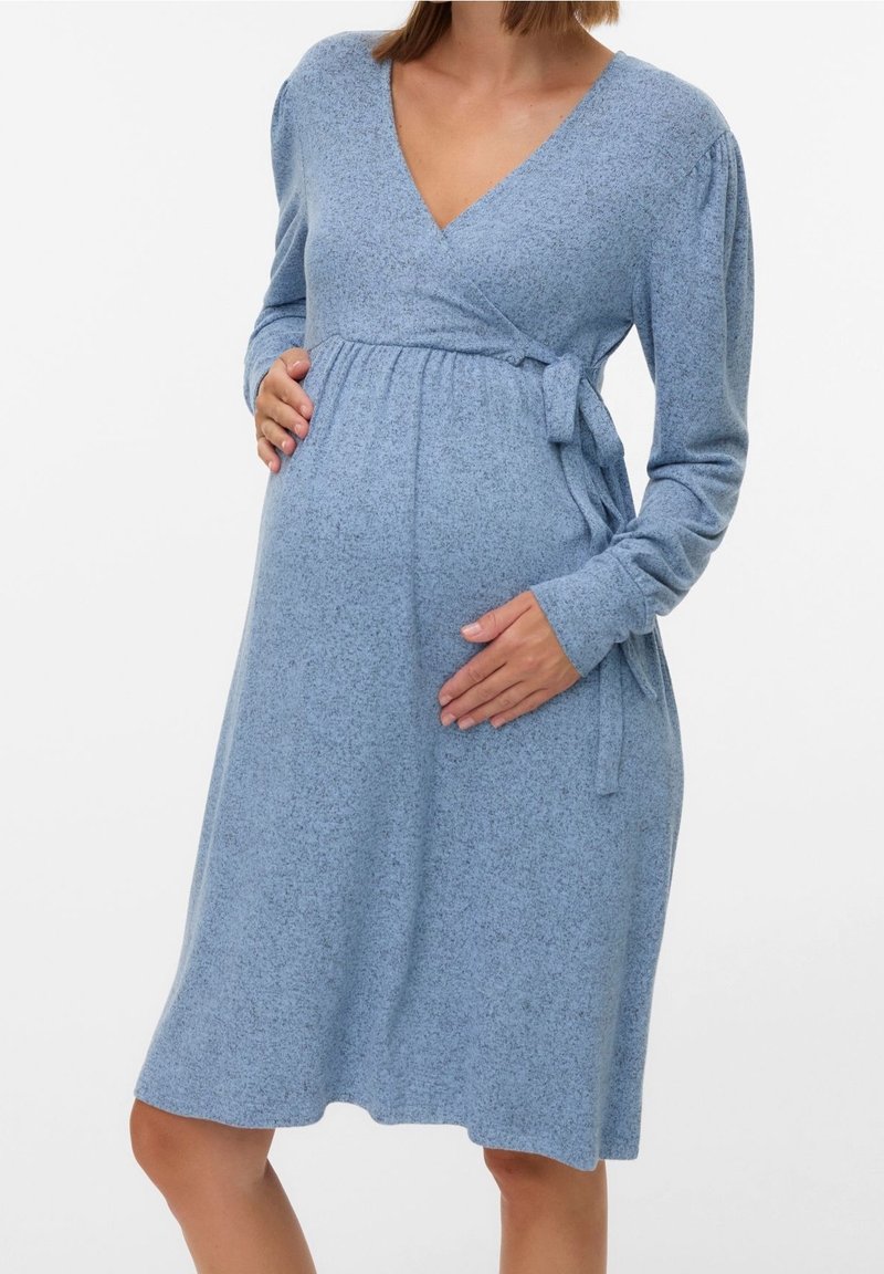 Femme enceinte portant une robe portefeuille à manches longues bleu clair avec un lien à la taille, les mains reposant sur son ventre.