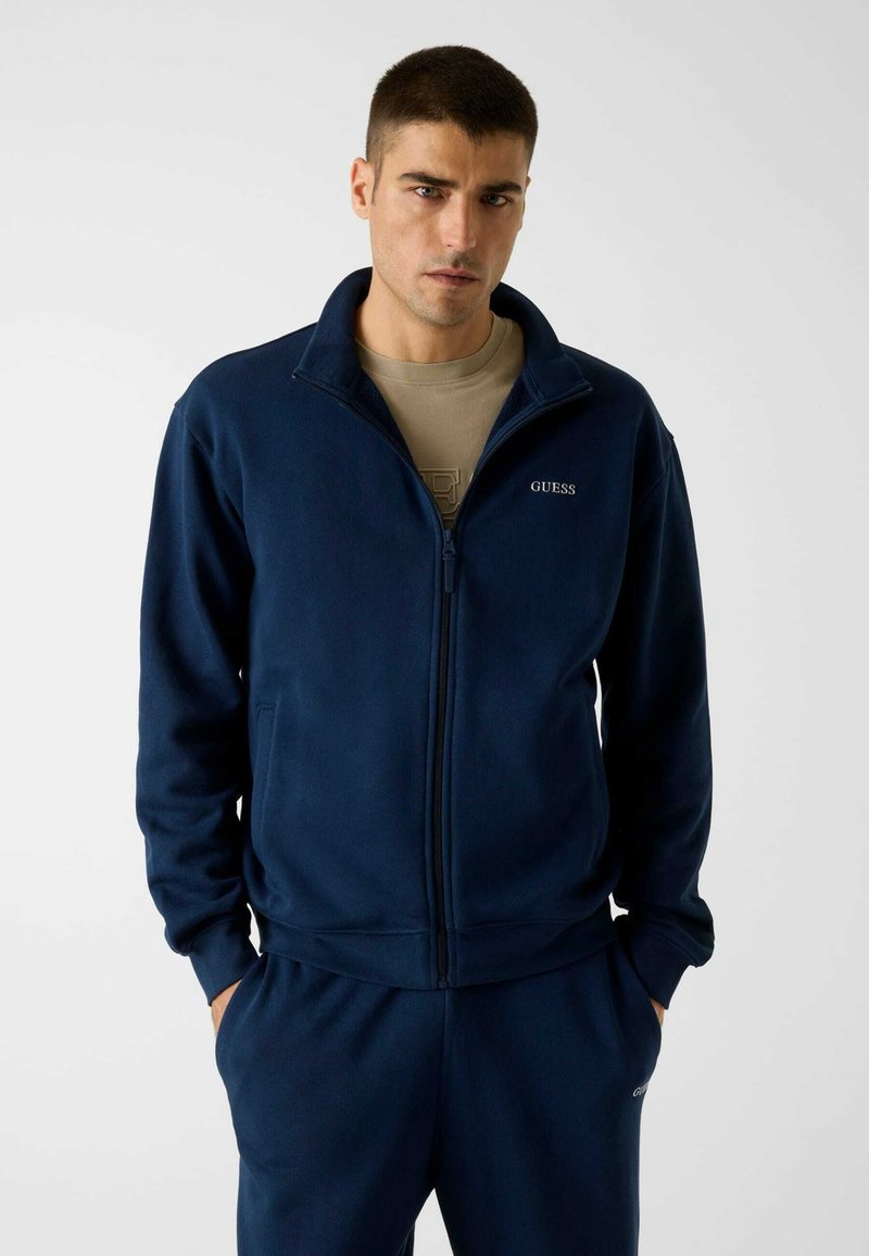 Veste polaire bleu marine à fermeture éclair avec un col montant, arborant un logo "GUESS" brodé sur la poitrine gauche et des poches latérales.