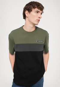 Tricou pentru bărbați din bumbac, cu un design colorat în verde oliv, gri și negru, cu guler rotund și mâneci scurte.