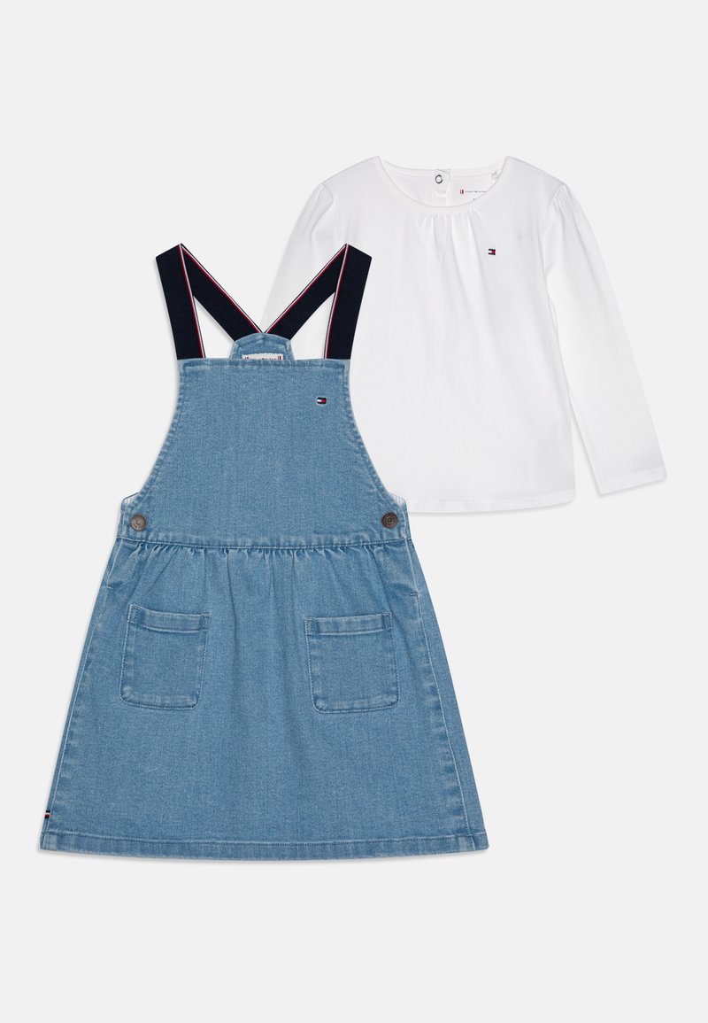 Conjunto de dos piezas de denim que incluye un vestido de tirantes azul con correas ajustables en color marino y dos bolsillos, combinado con una camisa de manga larga blanca.