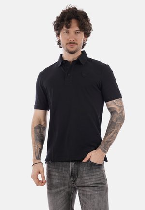 Uomo con capelli ricci che indossa una polo nera e jeans grigi, che mostra tatuaggi dettagliati su entrambi gli avambracci, in piedi contro uno sfondo bianco.