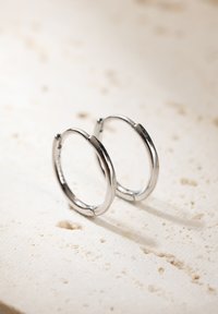 Pendientes de aro de plata con una superficie lisa y pulida, forma circular y bisagra reforzada para un cierre seguro, descansando sobre un fondo texturizado.