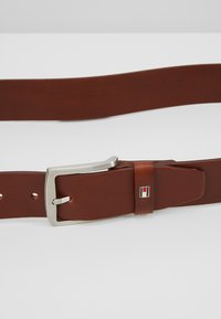 Ceinture en cuir marron avec une boucle en métal argenté et un petit logo en accent. Elle présente une texture lisse et plusieurs trous d'ajustement.