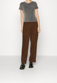 Szara, krótka koszulka z krótkim rękawem o wyblakłej teksturze i małym białym detalem tekstowym. Brązowe, luźne spodnie o prostym kroju. Czarne buty.