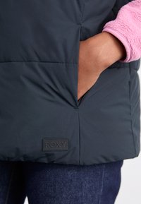 Giacca puffer di un blu scuro con un polsino in fleece rosa, dotata di tasca laterale e di un'etichetta nera con il logo Roxy all'orlo.