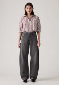 Sieviete, kas valkā sarkanas un baltas svītrām rakstītu pogājamā krekla ar sarullētām piedurknēm, platajām melnajām džinsām un melnām slip-on kurpēm, stāv uz vienkārša fona.