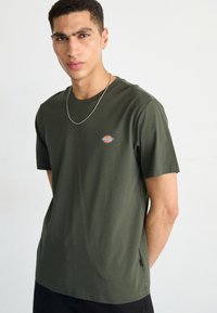 Tricou din bumbac verde măsliniu, cu mâneci scurte, având un mic logo multicolor pe piept și un detaliu cu etichetă pe lateral.