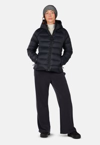 Giacca a vento nera con cappuccio, chiusura con zip e design trapuntato; abbinata a pantaloni scuri e a un berretto a righe bianco e nere.