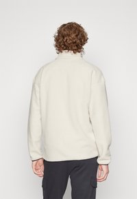 Pullover in felpa di colore bianco sporco con colletto alto, vestibilità over-size e tessuto testurizzato. Presenta spalle scese e una silhouette rilassata.