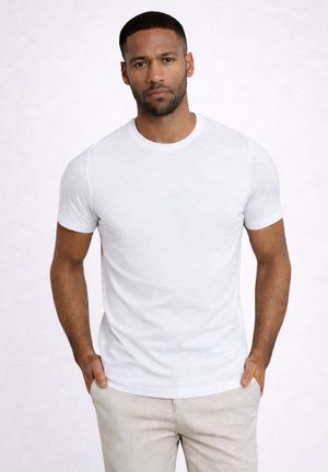 Mann mit kurzem Haar und Bart, der ein eng anliegendes, schlichtes weißes T-Shirt und hellbeige Hose trägt, mit den Händen in den Taschen vor hellem Hintergrund.