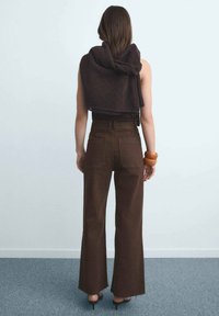 Pantalones marrones acampanados con bolsillos traseros y cintura alta, combinados con un chal oscuro texturizado drapeado sobre el hombro. Tacones negros visibles.