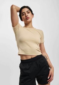 Person med kort mørkt hår iført en beiget avkortet skjorte og svarte shorts, poserer med en arm hevet og en synlig tatovering på underarmen.